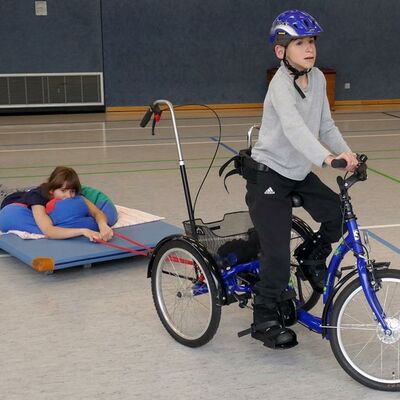 Schüler zieht mit Therapierad Schülerin auf Rollbrett und Matte liegend durch die Turnhalle.